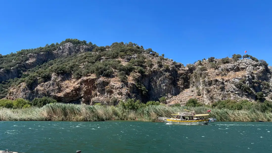 Dalyan Çayı üzerinde sarı tenteli bir tur teknesi; kıyıda sazlıklar, arkada Kaunos kayalıkları